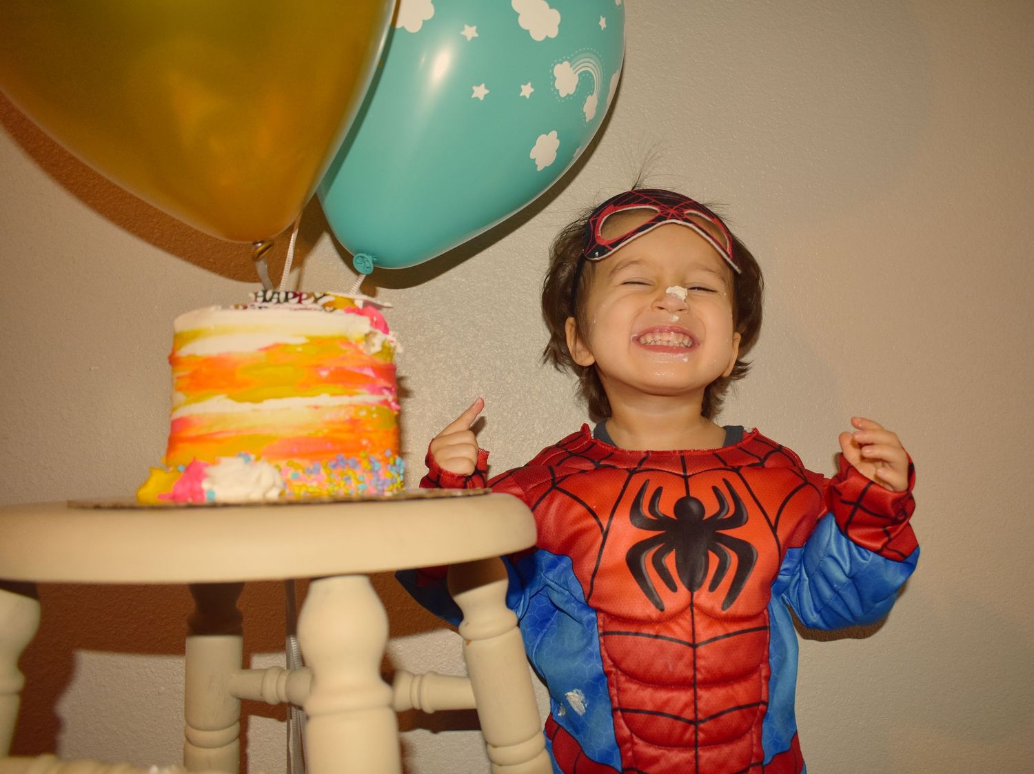 A Spider-Man costume stands next to a colorful birthday cake with orange frosting and festive balloons.