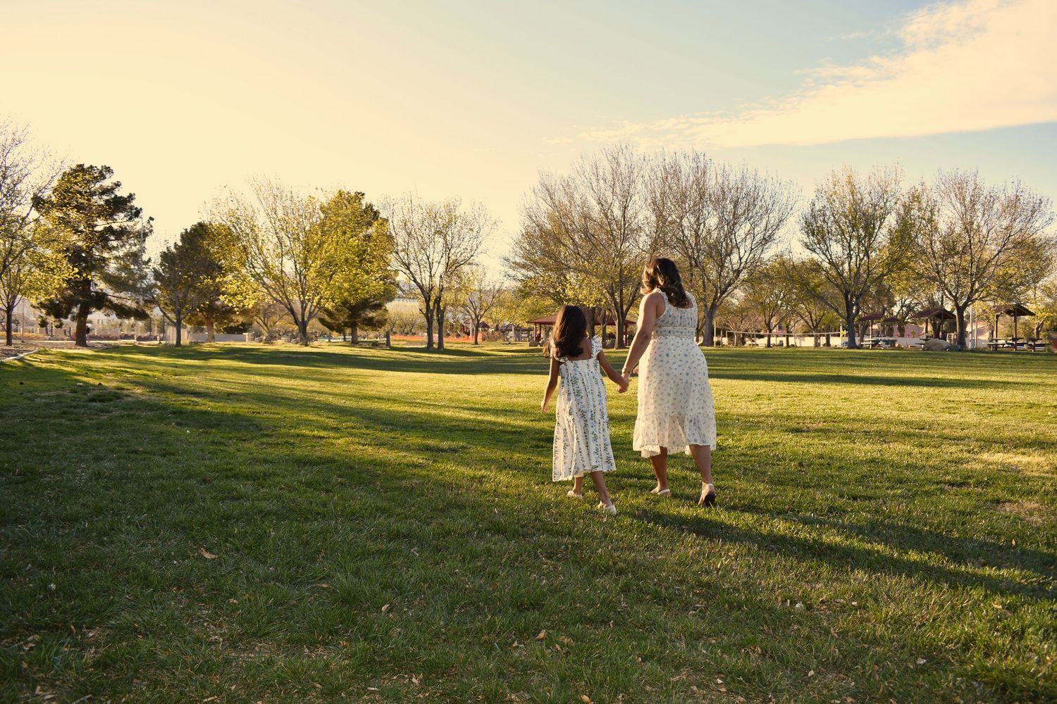 Two girls in white dresses hold hands walking through a sunny green park with bare trees in the background.