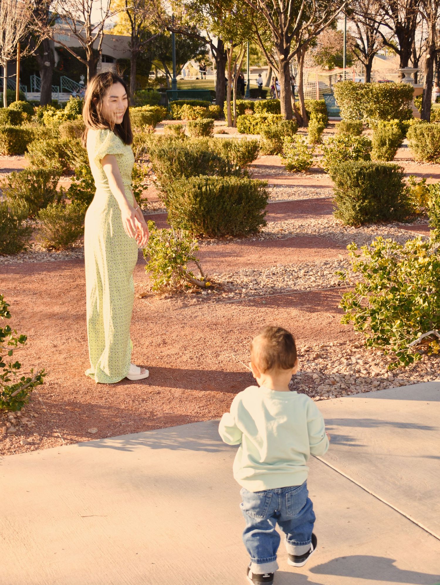 A toddler in jeans and light sweater walks along a paved path surrounded by desert landscaping and shrubs.