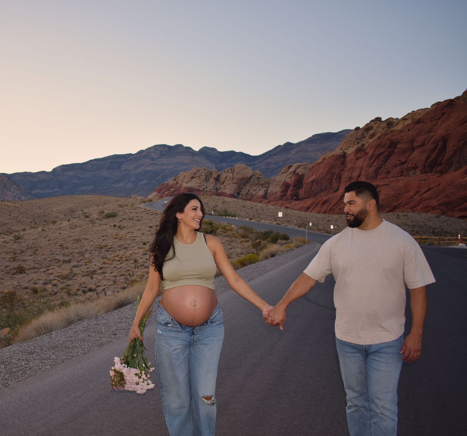 A couple holds hands while walking on a desert road with red rock cliffs in the background.