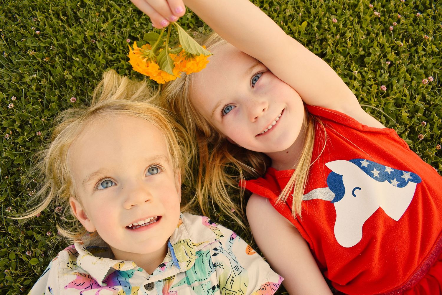 Two children lying on grass with flowers, wearing colorful summer outfits.