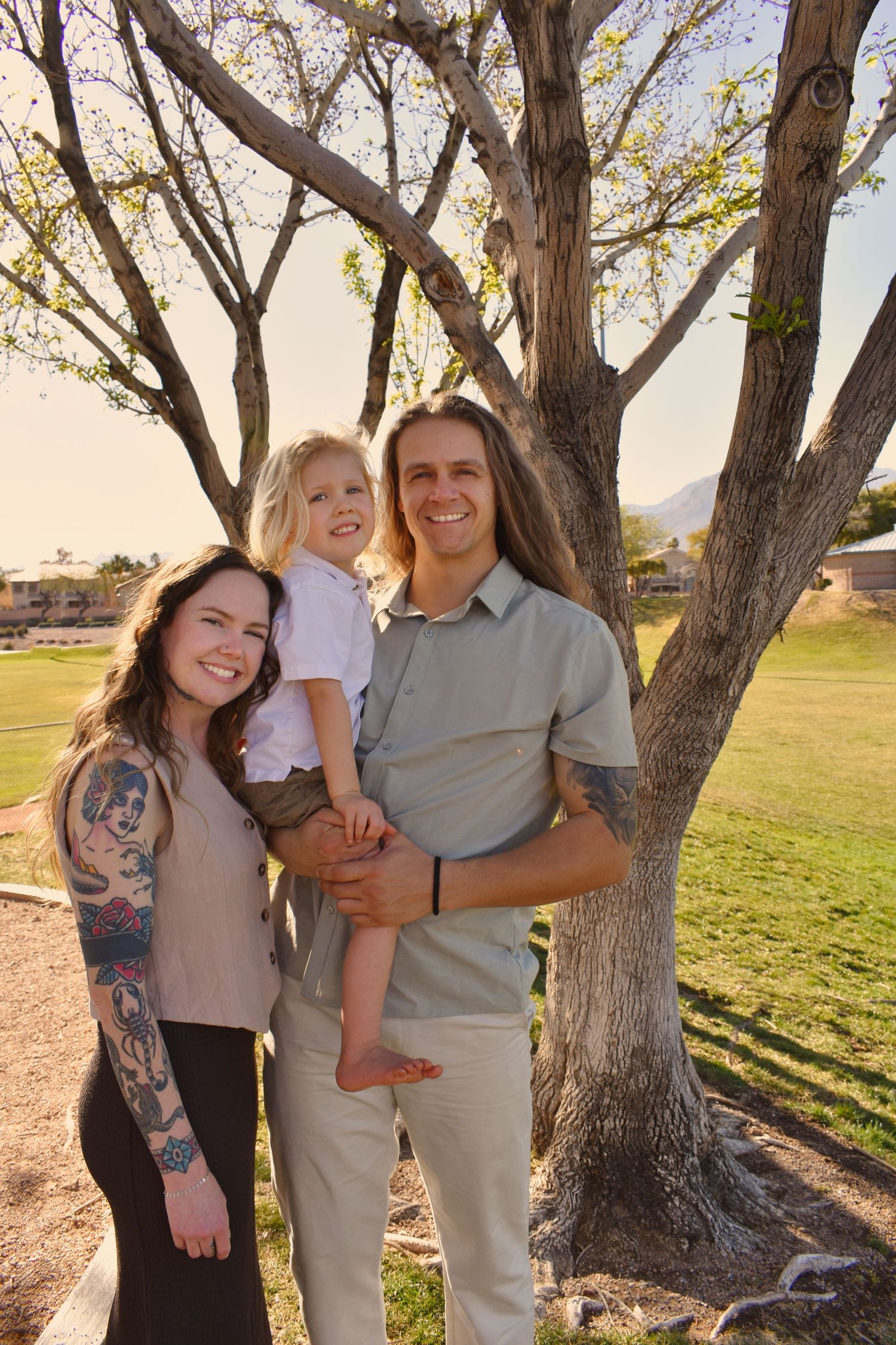 A family poses together under large trees in a park during golden hour lighting.