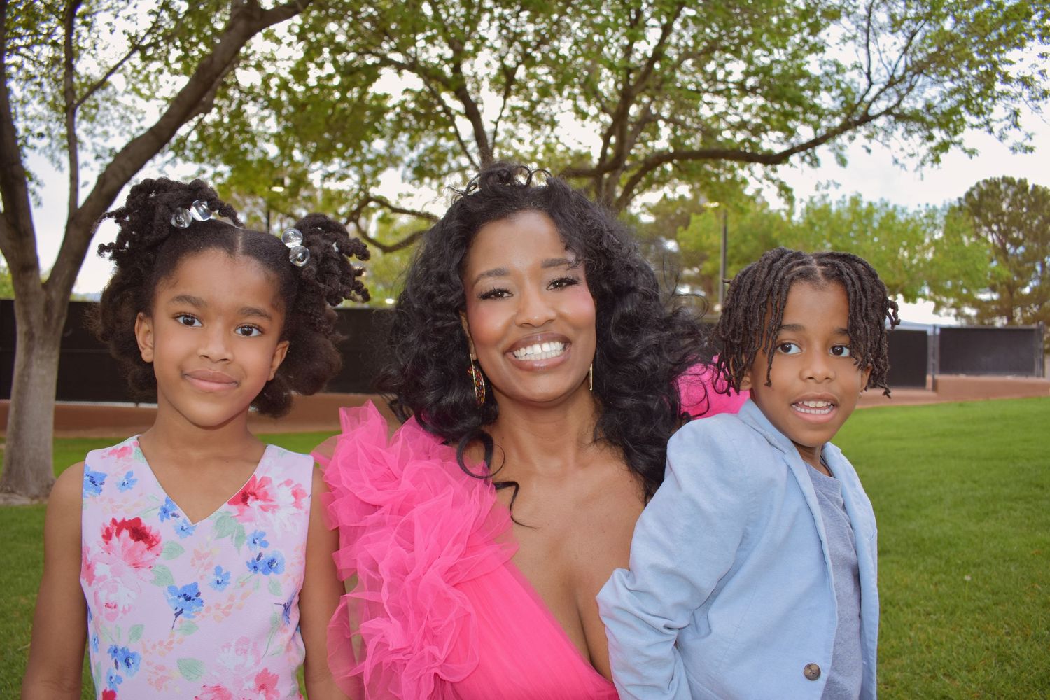 A smiling woman in a pink ruffled dress poses outdoors with two young children in pastel outfits near green trees.