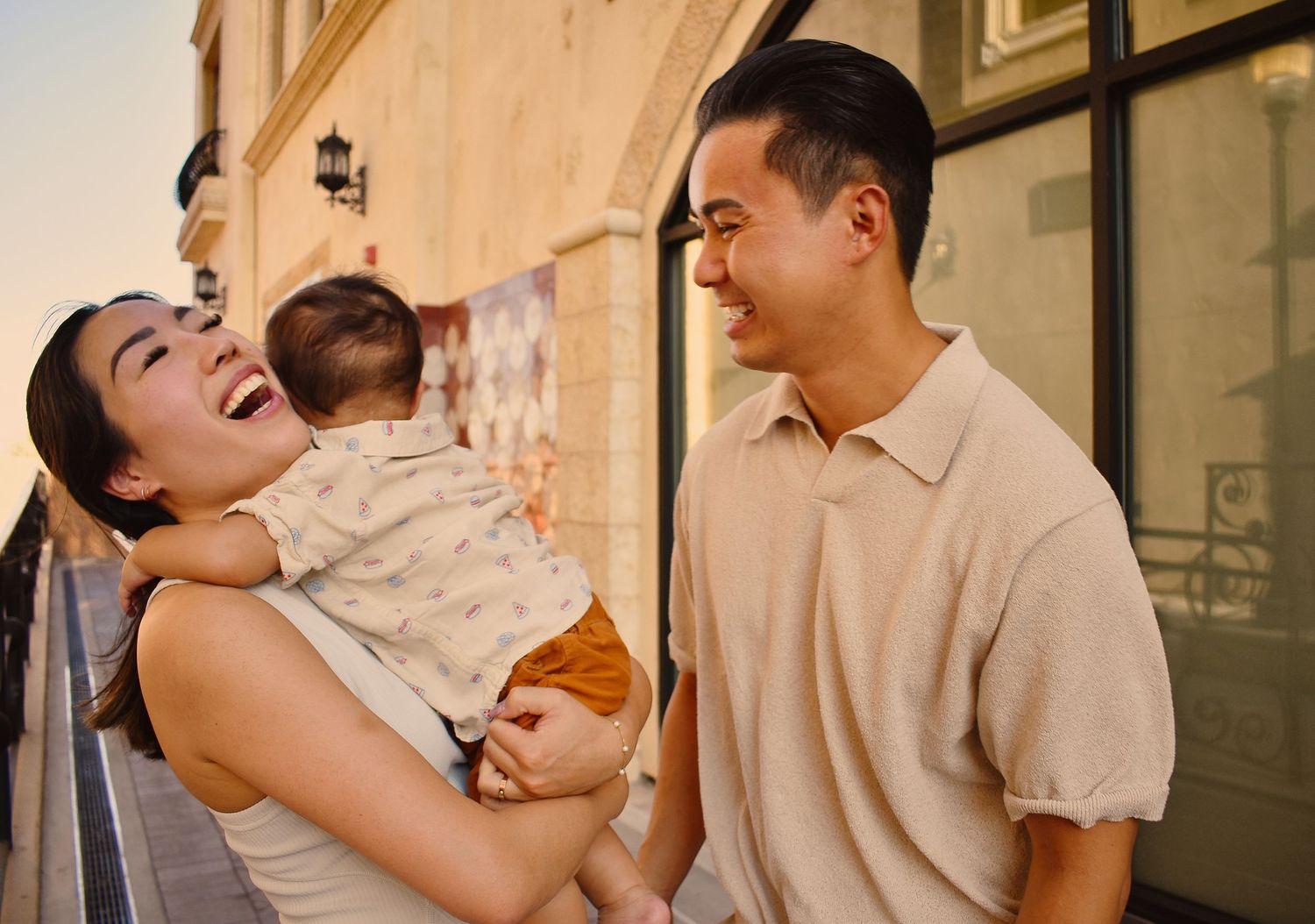 A family laughs and plays together in front of a neutral-colored building exterior.