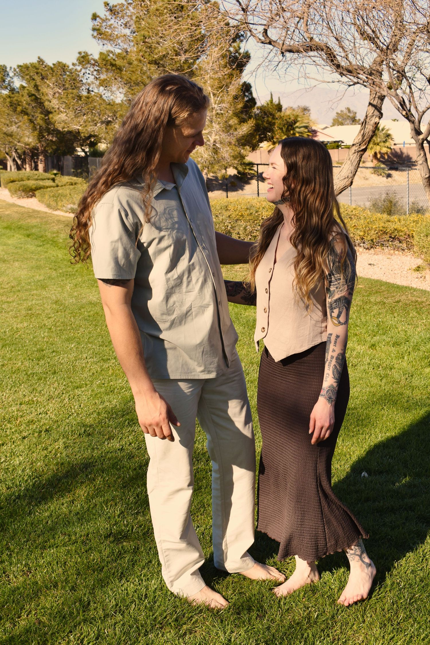 Two people in casual attire standing together on green grass in a sunny park setting.