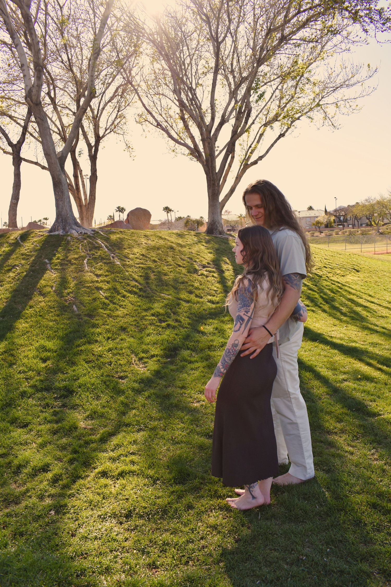 A couple shares a tender moment on a grassy hillside at sunset with long shadows cast by trees in the background.
