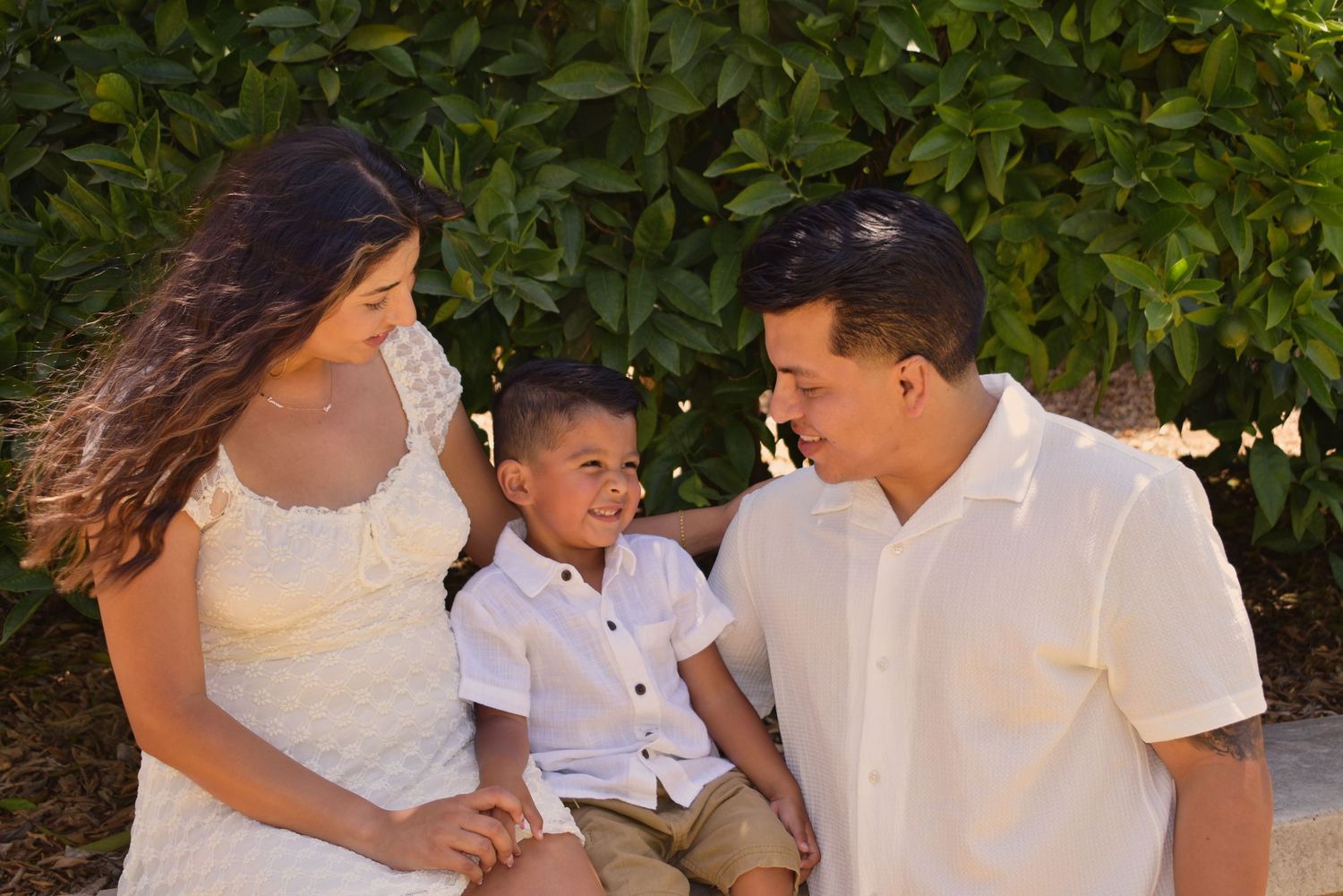 Family in white outfits sitting together outdoors against a leafy green background.