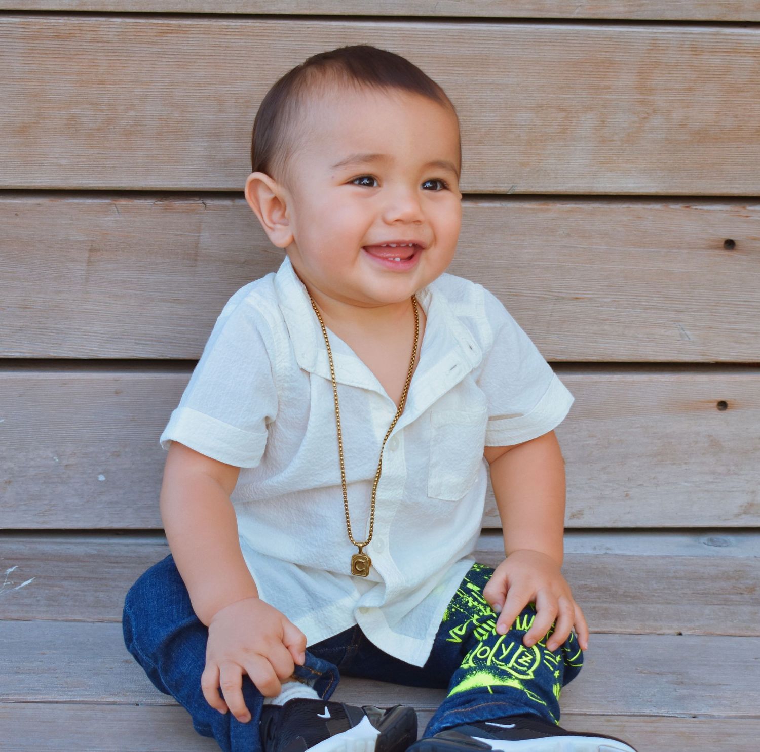 A toddler in a white shirt and blue jeans sits on wooden steps with a cheerful expression.
