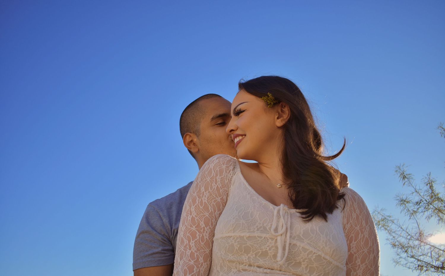 A romantic couple embraces outdoors against a bright blue sky during a photo session.