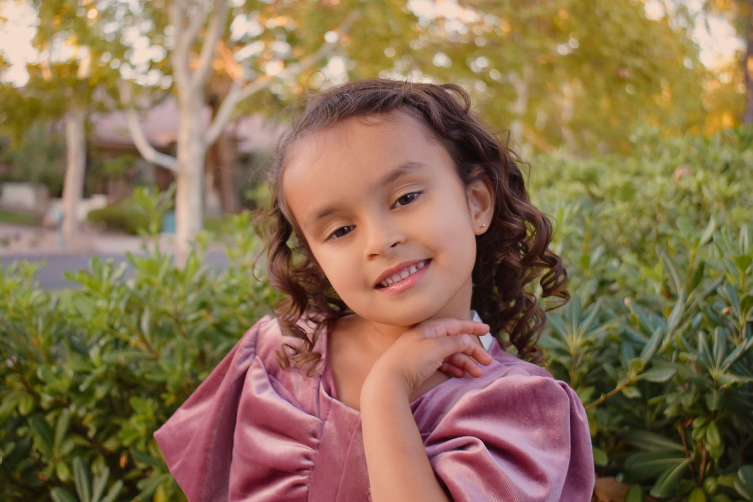 Child in pink dress poses outdoors against green foliage background.