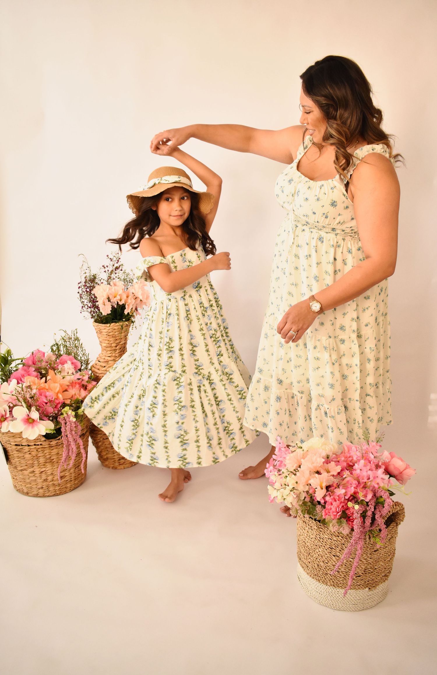 Mother and daughter in matching floral maxi dresses pose with wicker baskets of pink flowers in a bright studio.