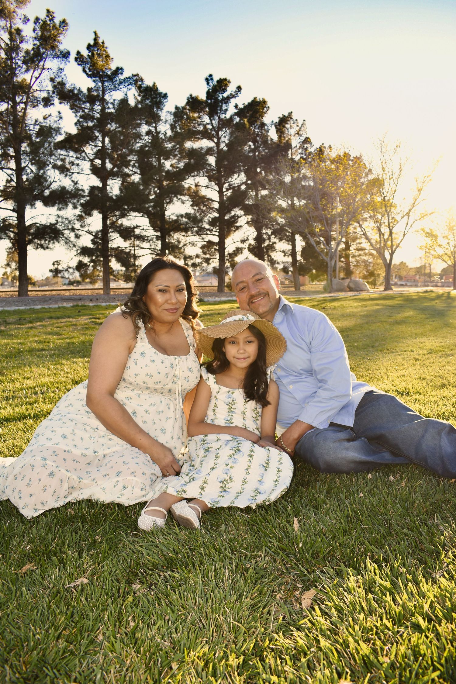 Family of three sitting on green grass in a sunny park, parents smiling with young girl in straw hat between them.