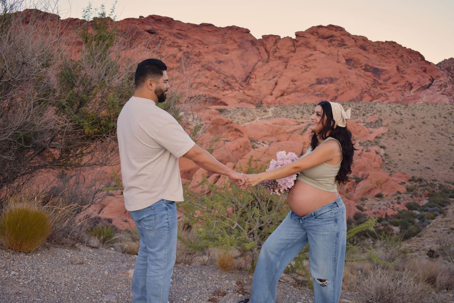 A couple stands together against a backdrop of red desert cliffs during golden hour.