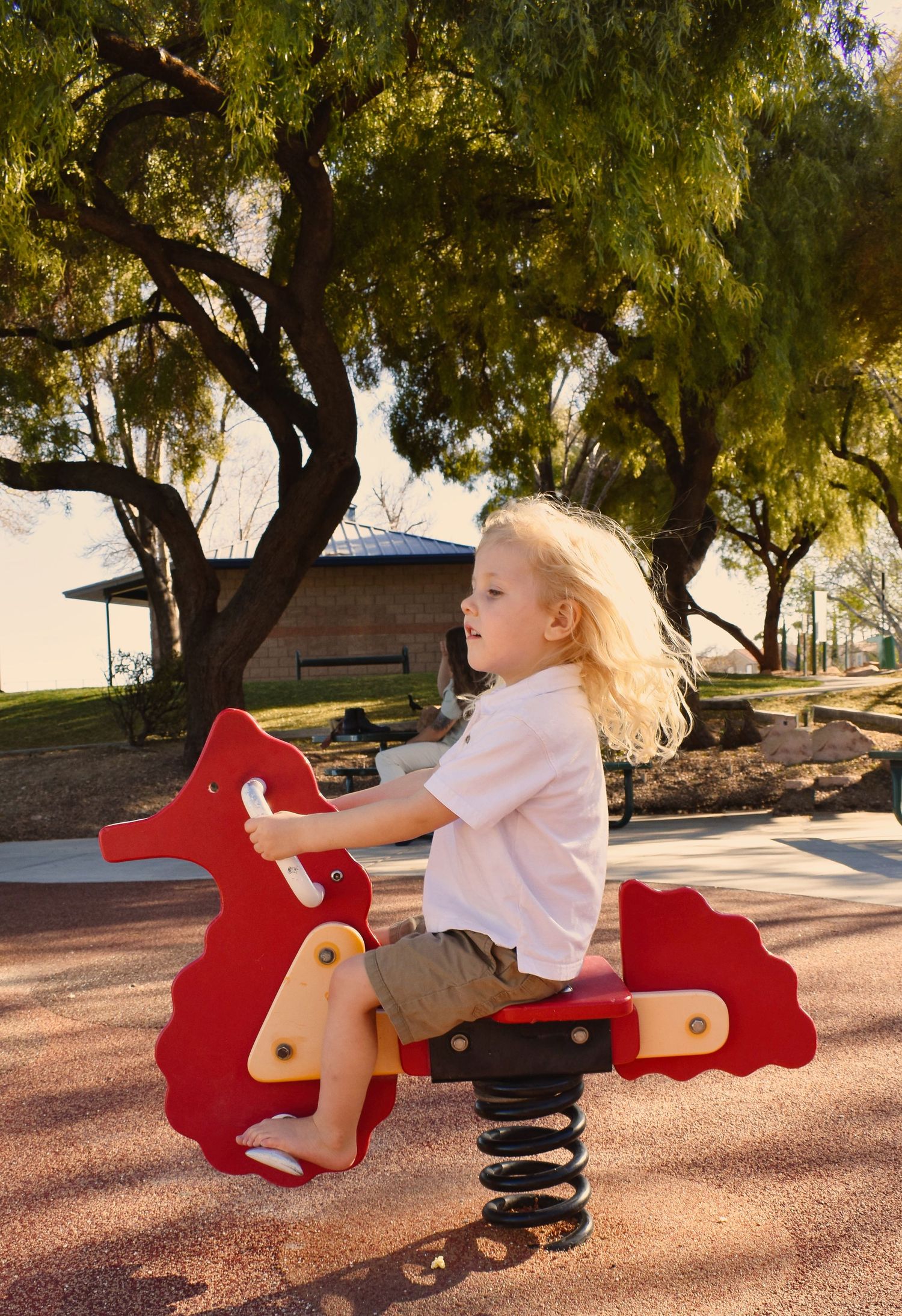 A child plays on a red spring rider toy in a park with trees visible in the background.