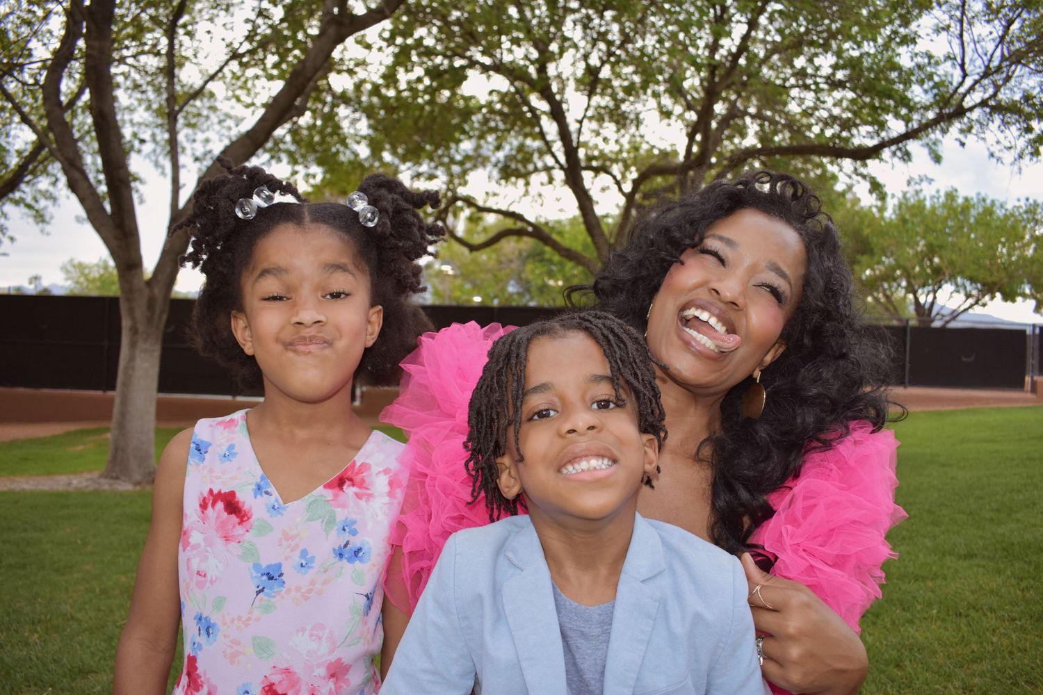 A smiling Black woman in a pink tutu poses outdoors with two young children in a green park setting.