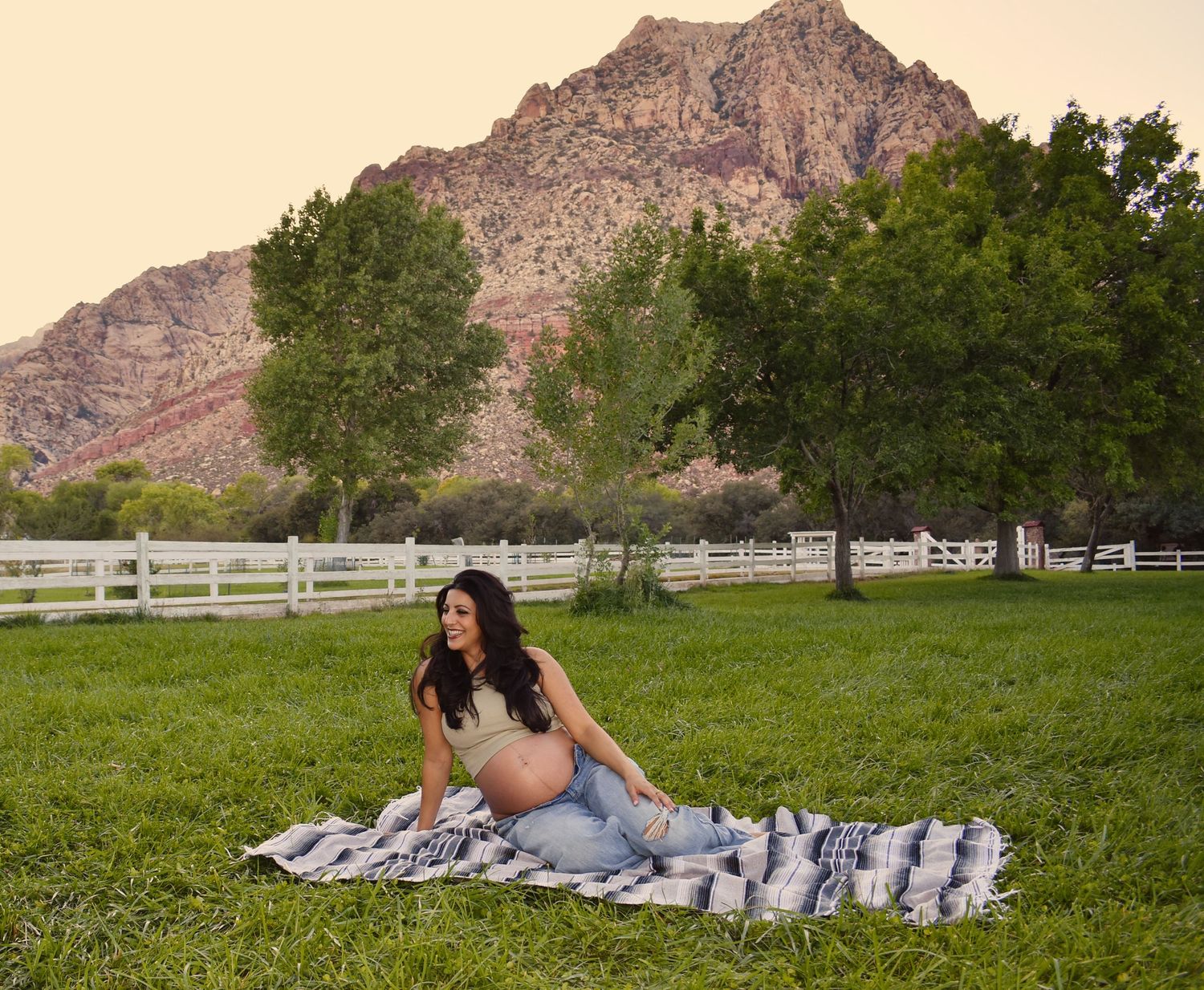 A maternity photo shoot on a picnic blanket with mountains and white fence in background.
