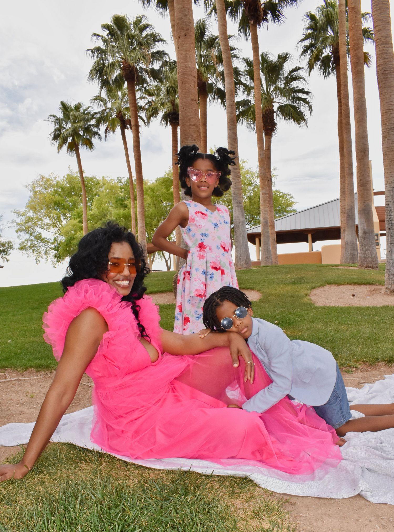 A pregnant woman in a pink dress poses with two children on grass, surrounded by tall palm trees in a sunny park.
