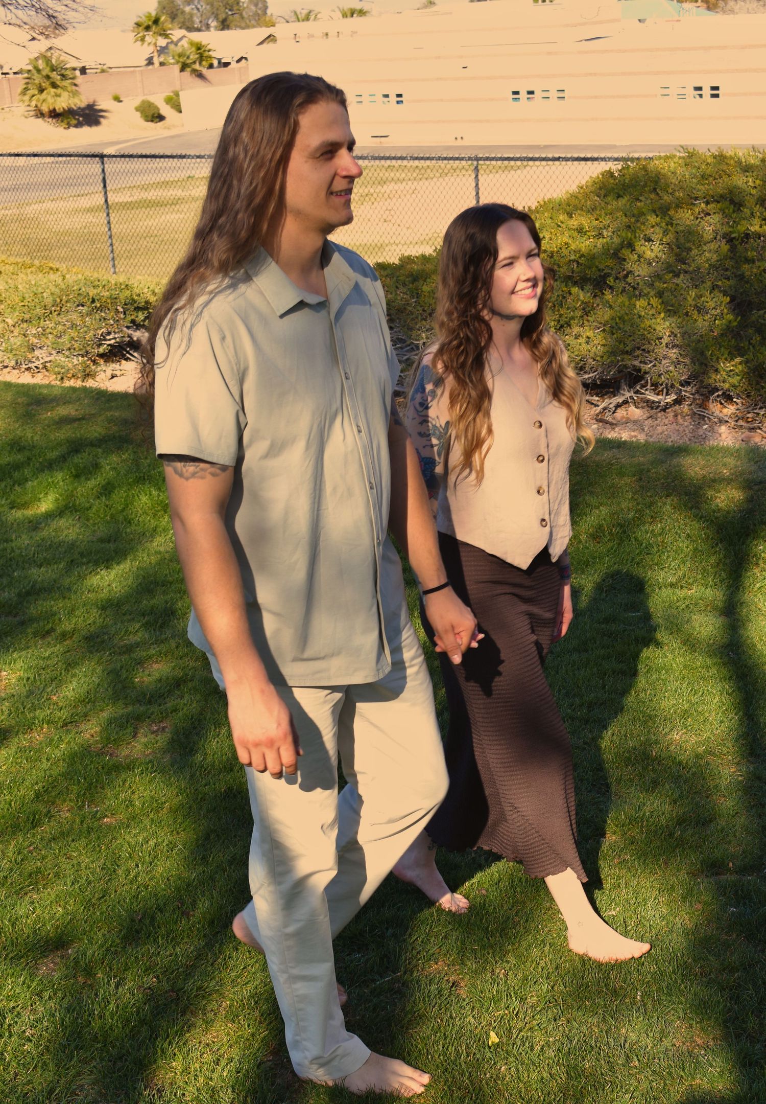 A couple stands together on a green lawn at sunset, wearing casual attire and holding hands.