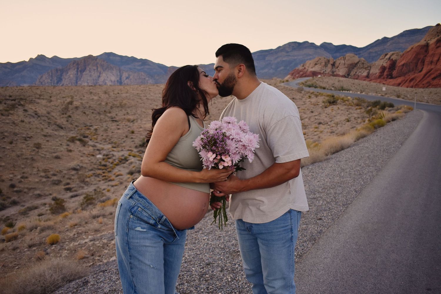 A couple shares a tender moment on a desert road with mountains in the background.