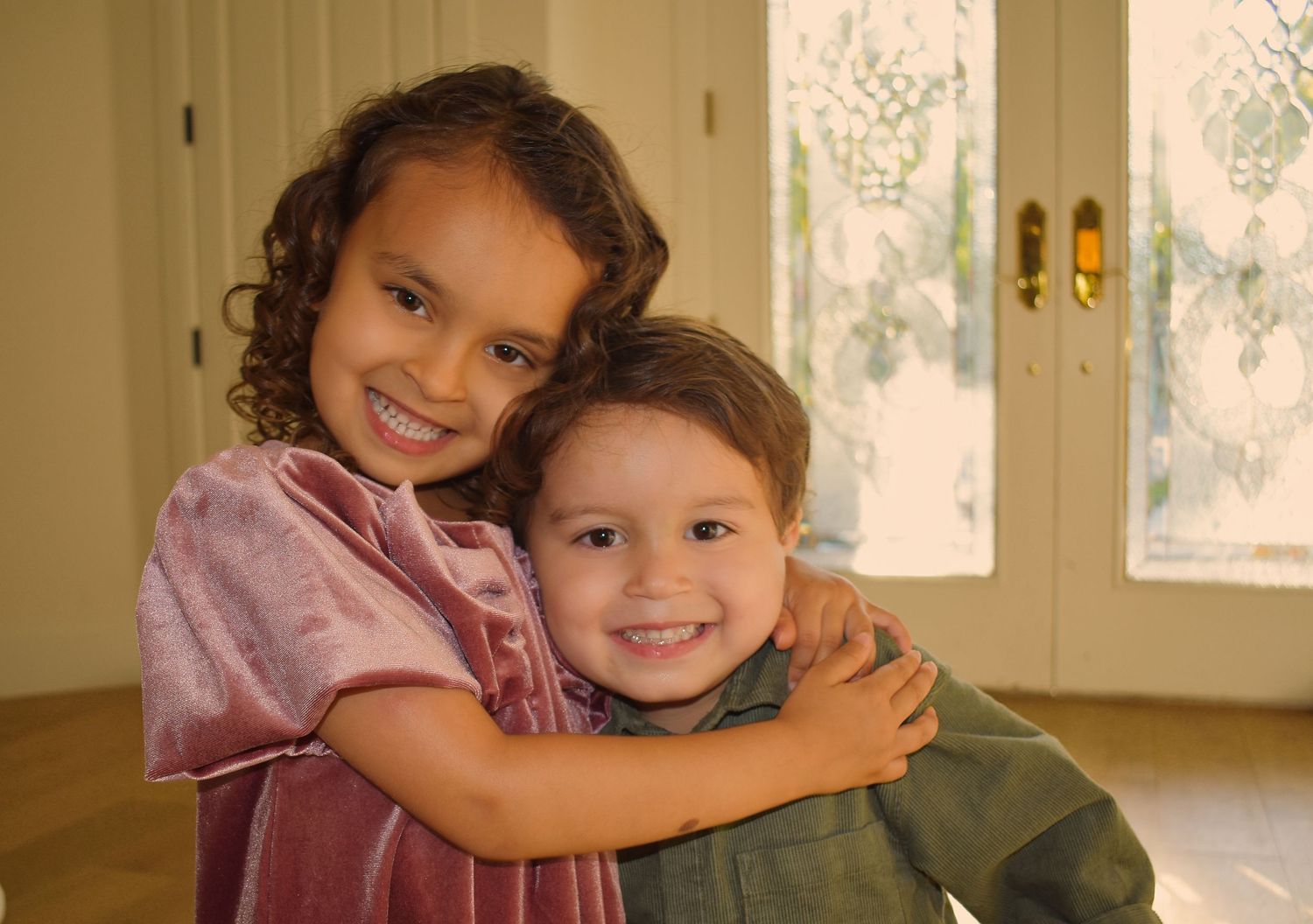 Two children share a warm embrace while smiling in a sunlit room.