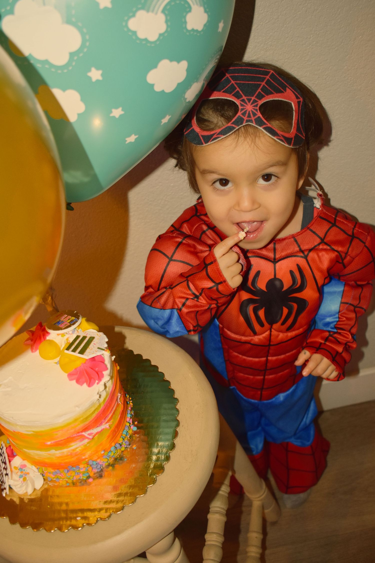 A Spider-Man costume and colorful balloons surround a festive birthday cake on a wooden table.