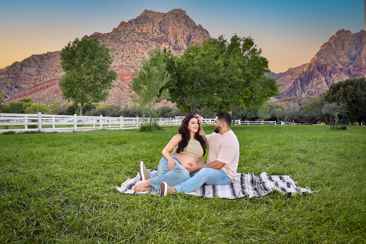 Couple having picnic on grass with mountain backdrop and white fence.