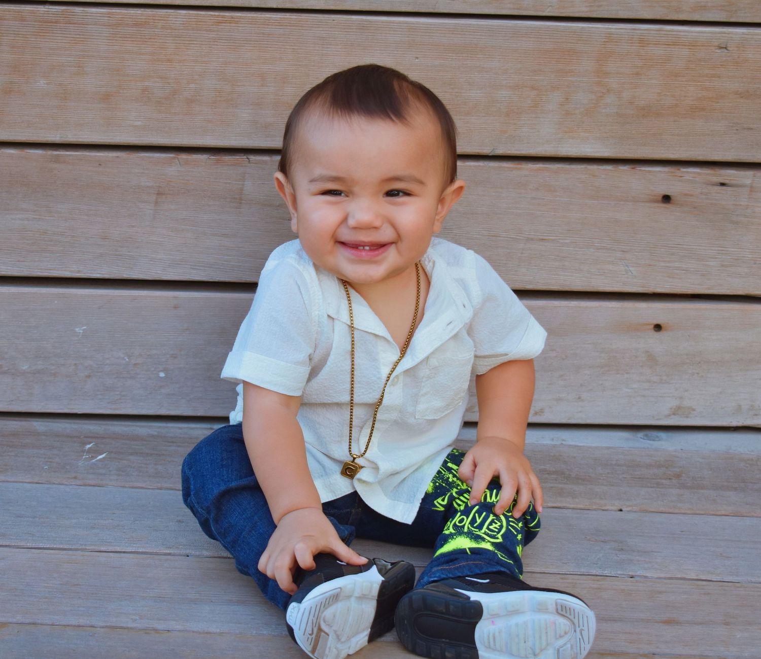 Baby in white outfit sitting on wooden deck in sunlight.