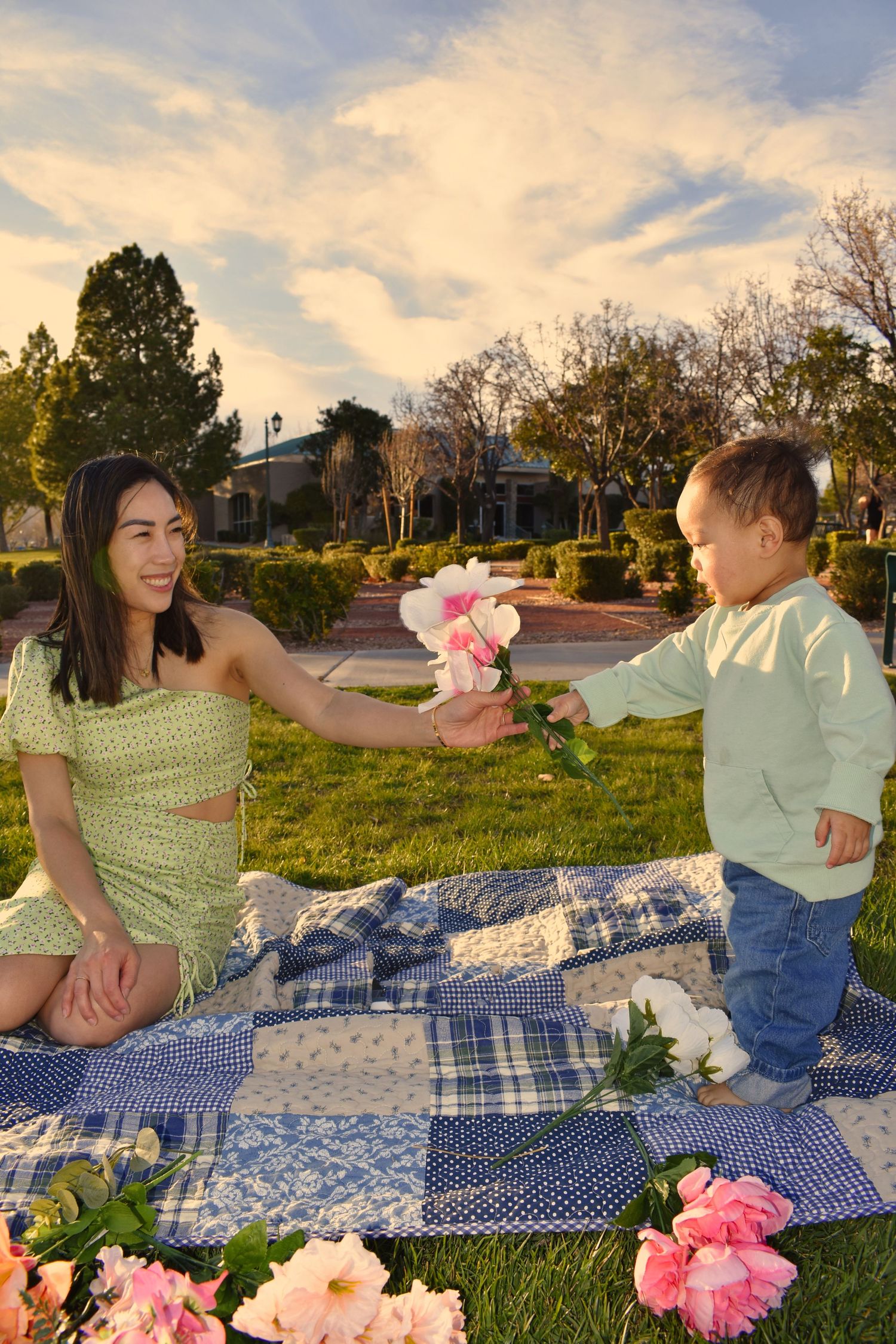 Two people sit on a blue checkered blanket in a grassy park at sunset with pink flowers nearby.