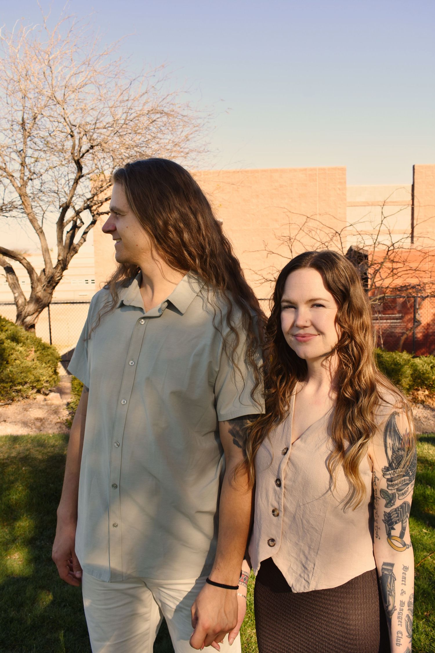 Two people stand together outdoors at sunset with cherry blossom trees in the background.