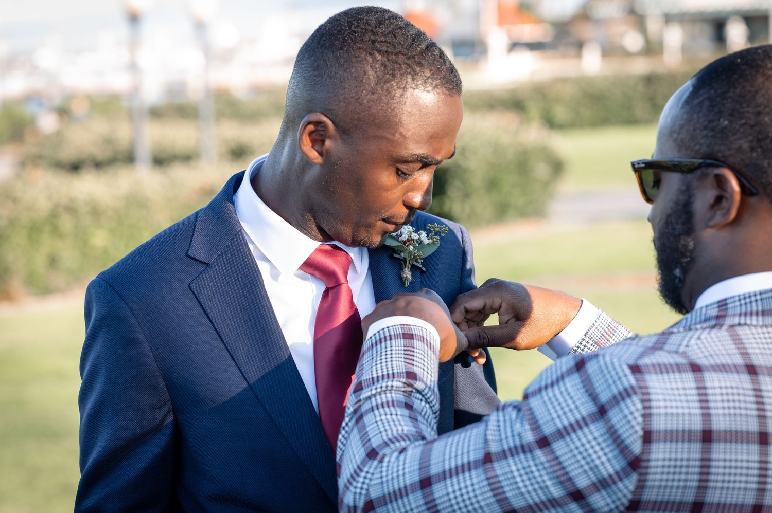 A groomsman helps adjust a boutonniere on the groom's navy suit jacket outdoors.