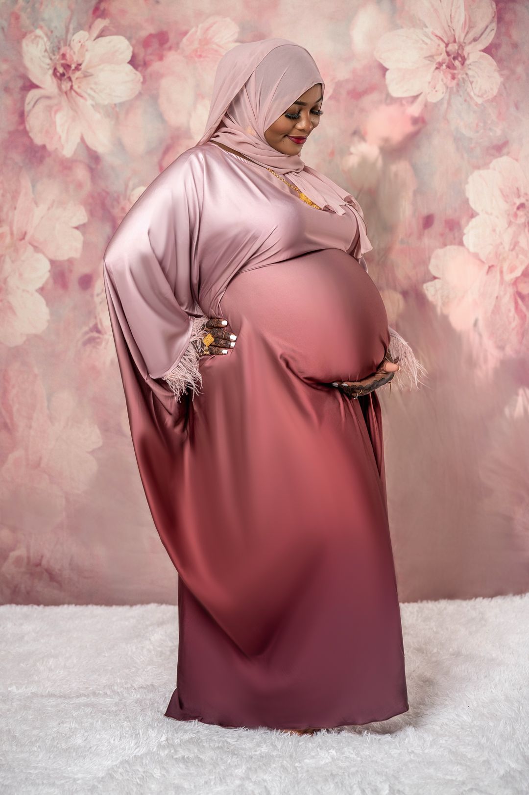 Pregnant woman in pink ombre dress and hijab poses against floral backdrop for maternity photo.