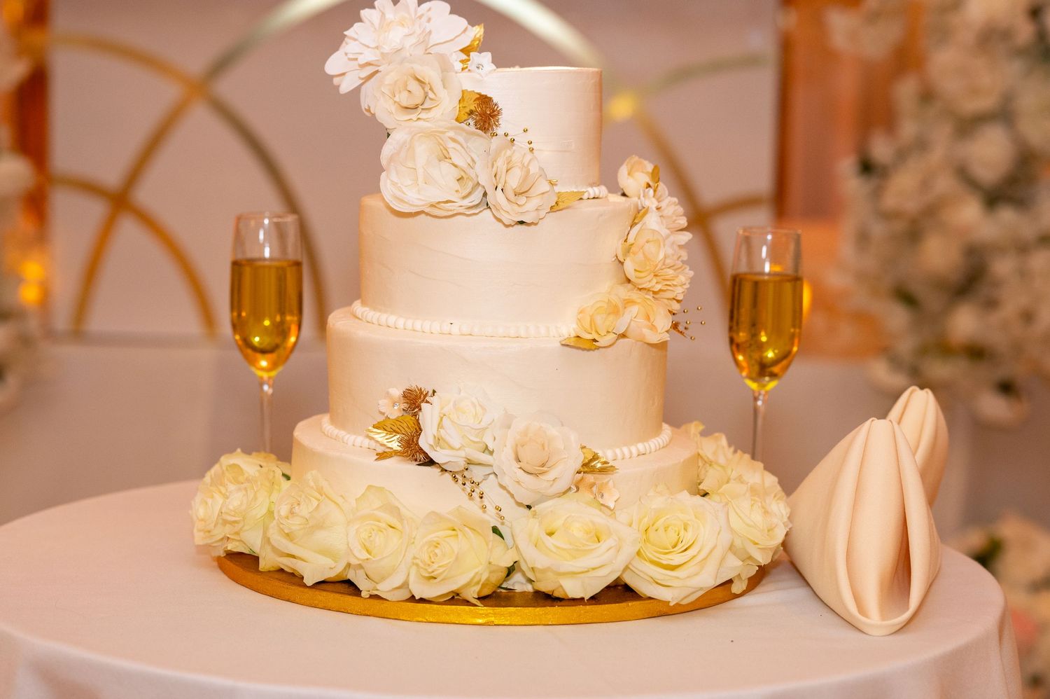 Elegant white wedding cake decorated with roses and gold cherubs, flanked by champagne flutes.