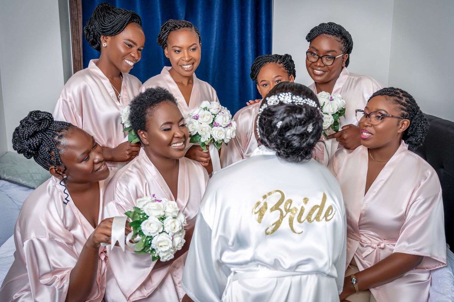 Bridesmaids in pink robes gather around bride wearing white robe during wedding morning preparations.