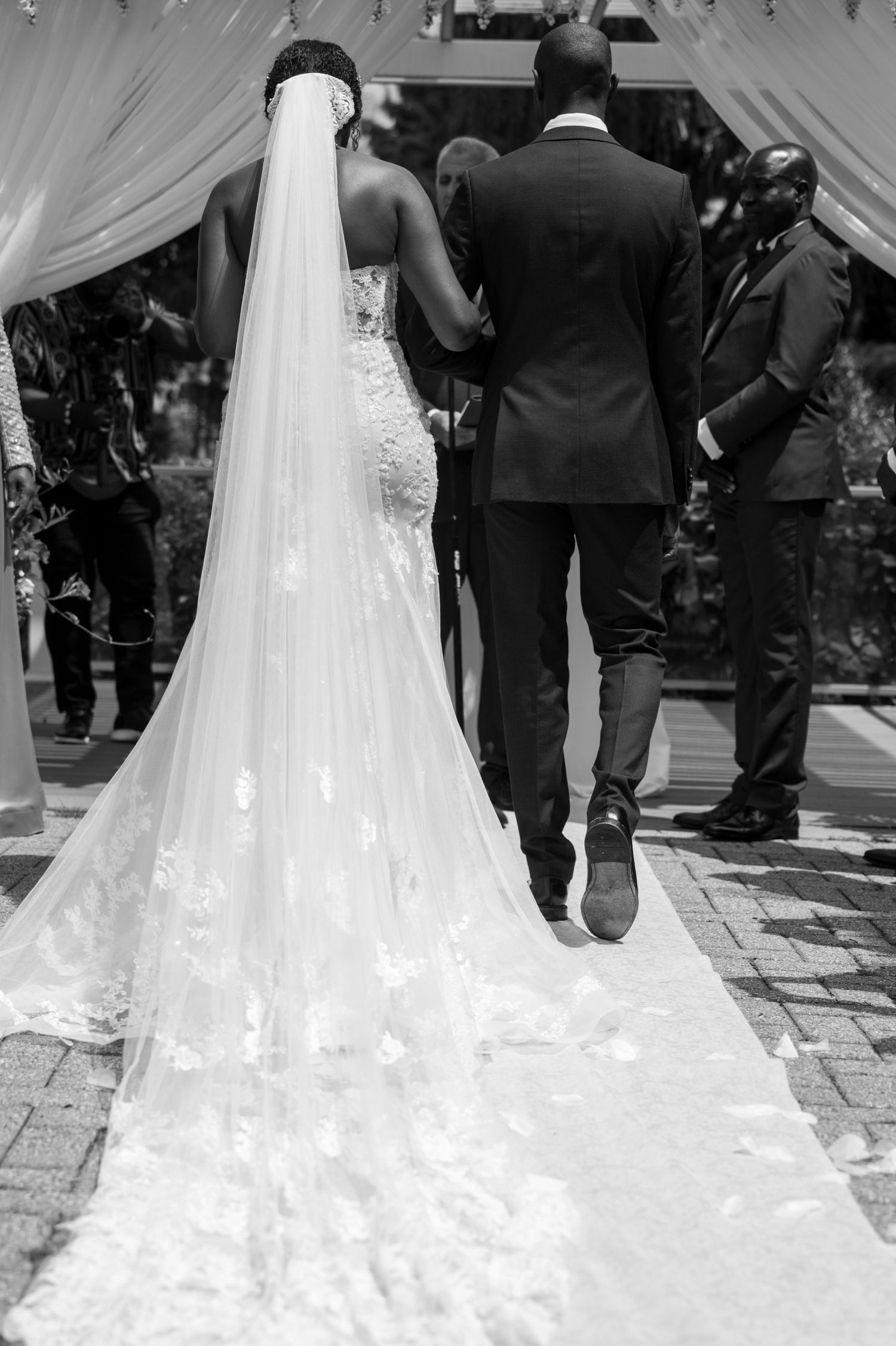 Black and white photo of couple walking under draped ceremony arch with flowing wedding dress train.