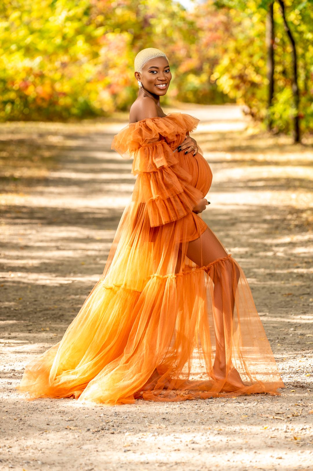 Pregnant woman in flowing orange tulle gown poses on autumn path during maternity session.
