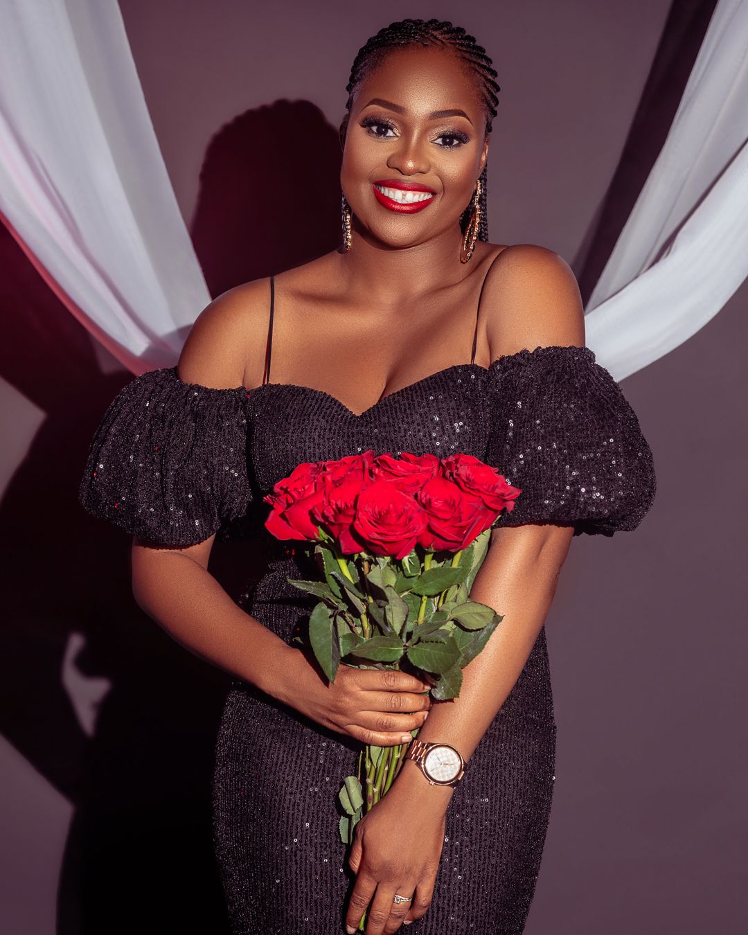 Elegant portrait in black sparkly off-shoulder dress holding red roses against draped background.