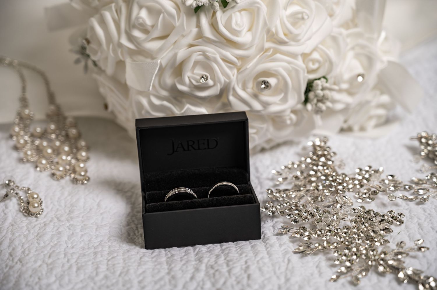Wedding rings nestled in a black box surrounded by white roses and pearl decorations.