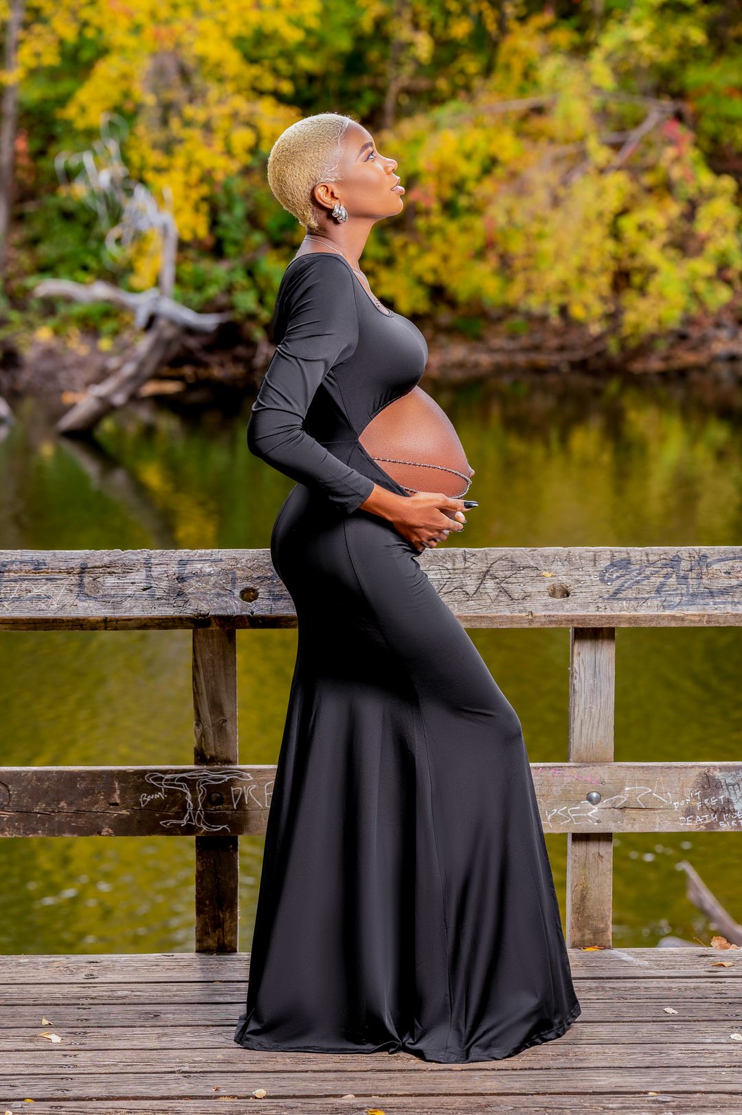 Pregnant woman in black fitted dress poses on wooden bridge during autumn photoshoot.