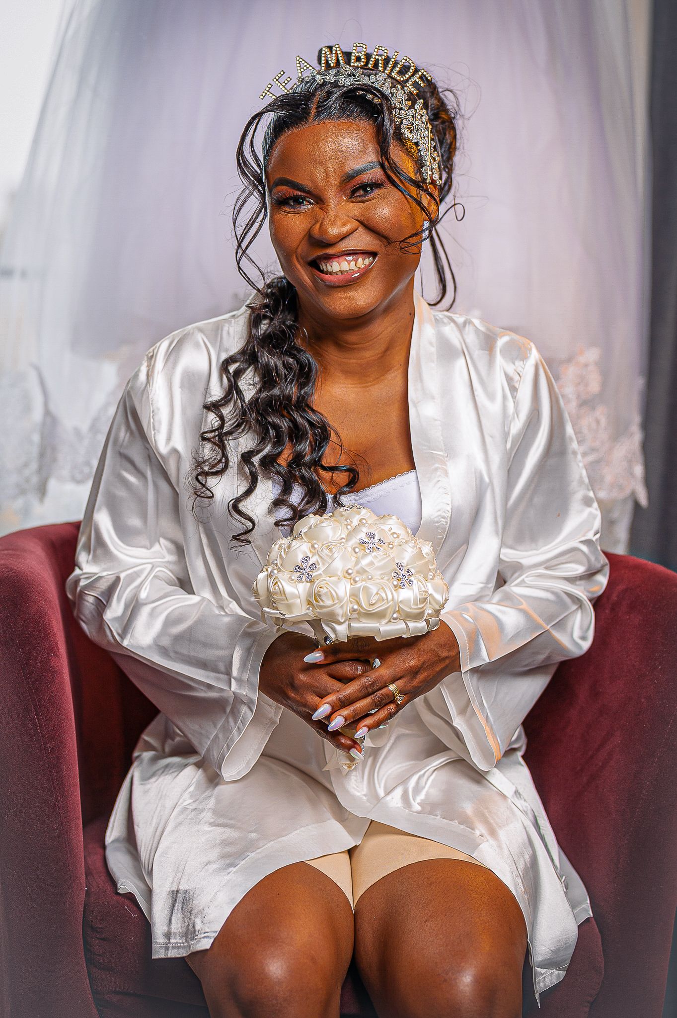 A radiant bride wearing a white silk robe and tiara during wedding preparations.