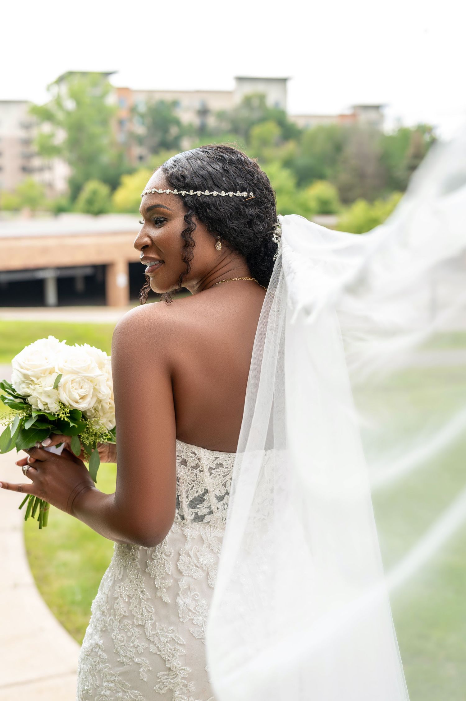 Side profile of bride in strapless gown with flowing veil and crystal headpiece holding white roses.