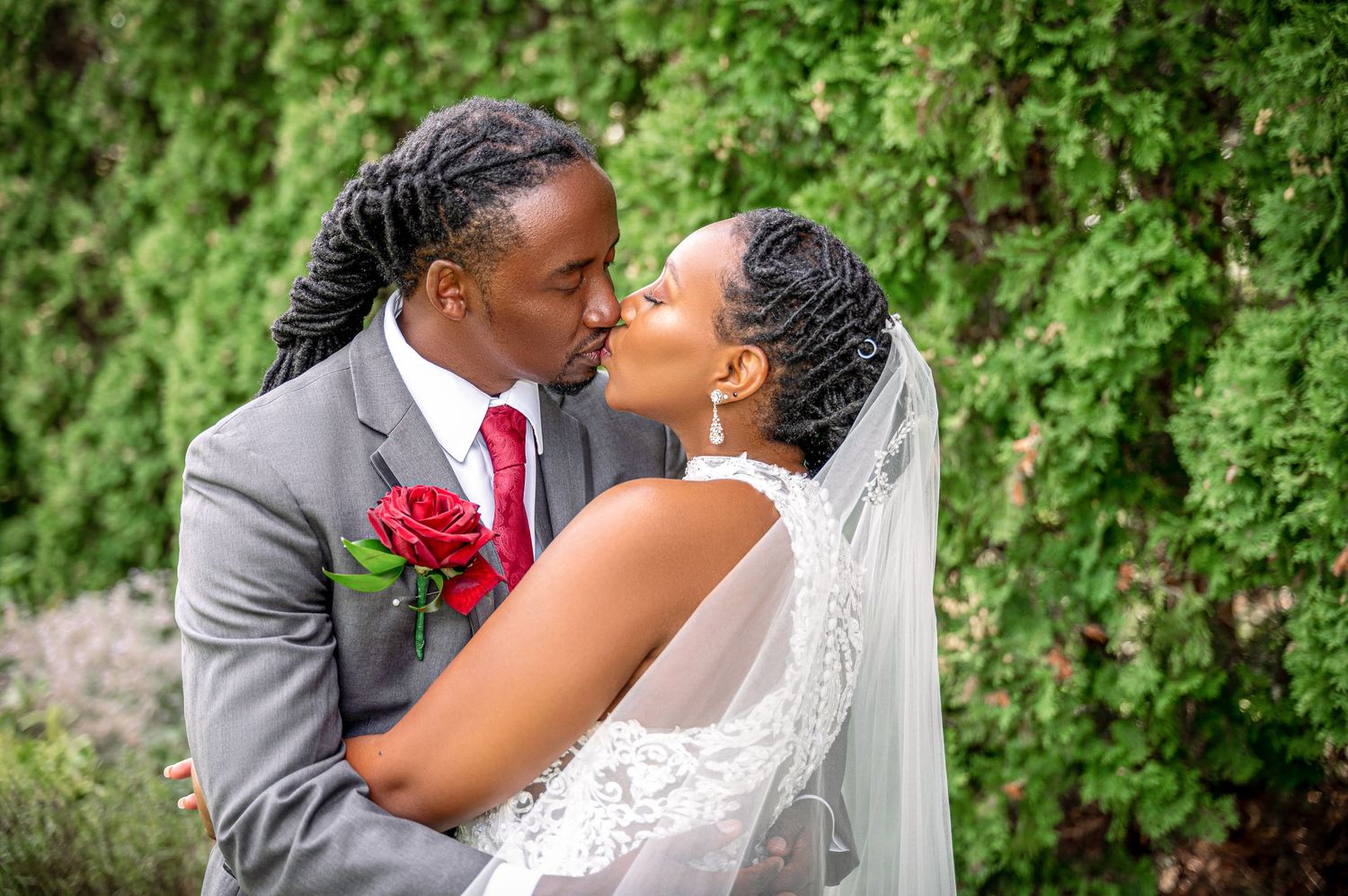 A romantic wedding portrait of a couple embracing in front of lush greenery with the groom wearing a red tie.