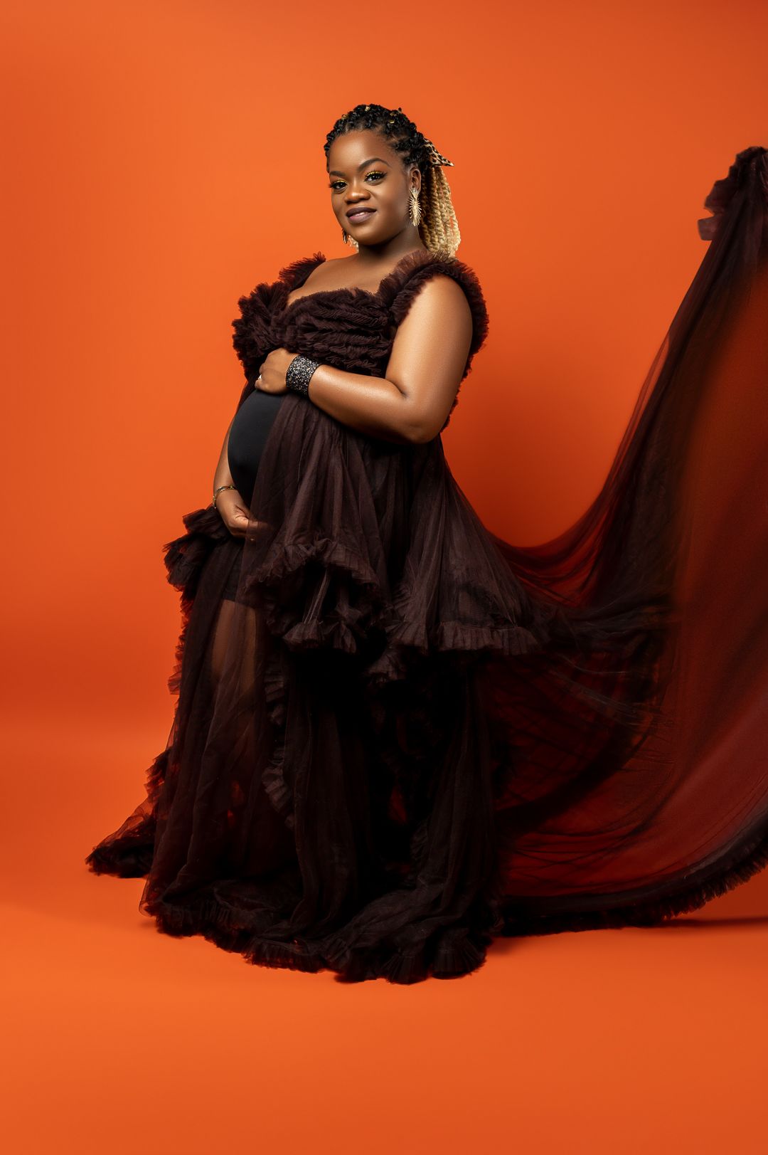Maternity portrait of woman in black ruffled gown against vibrant orange backdrop.