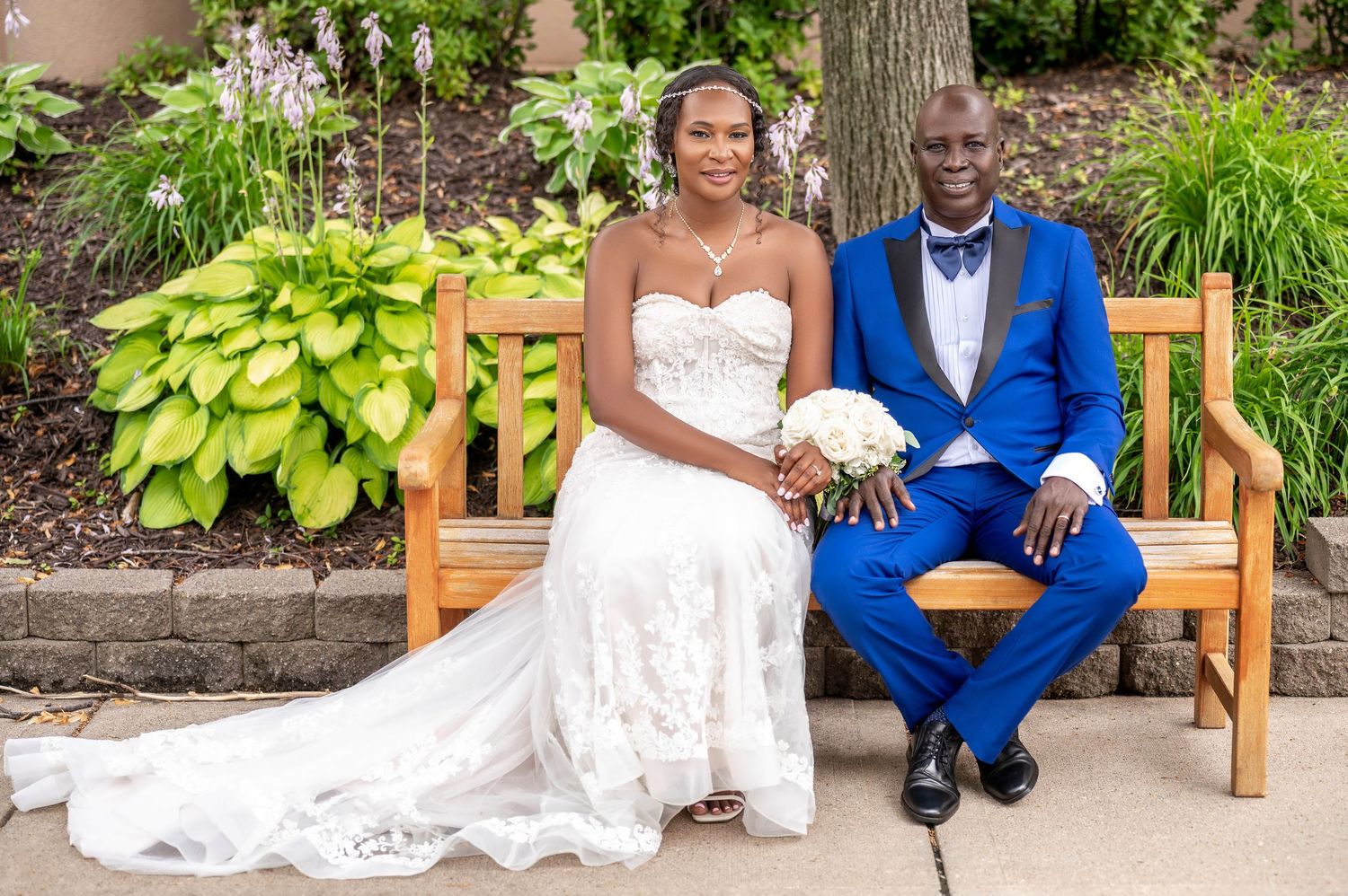 Wedding couple in elegant attire sitting on wooden garden bench surrounded by lush greenery.