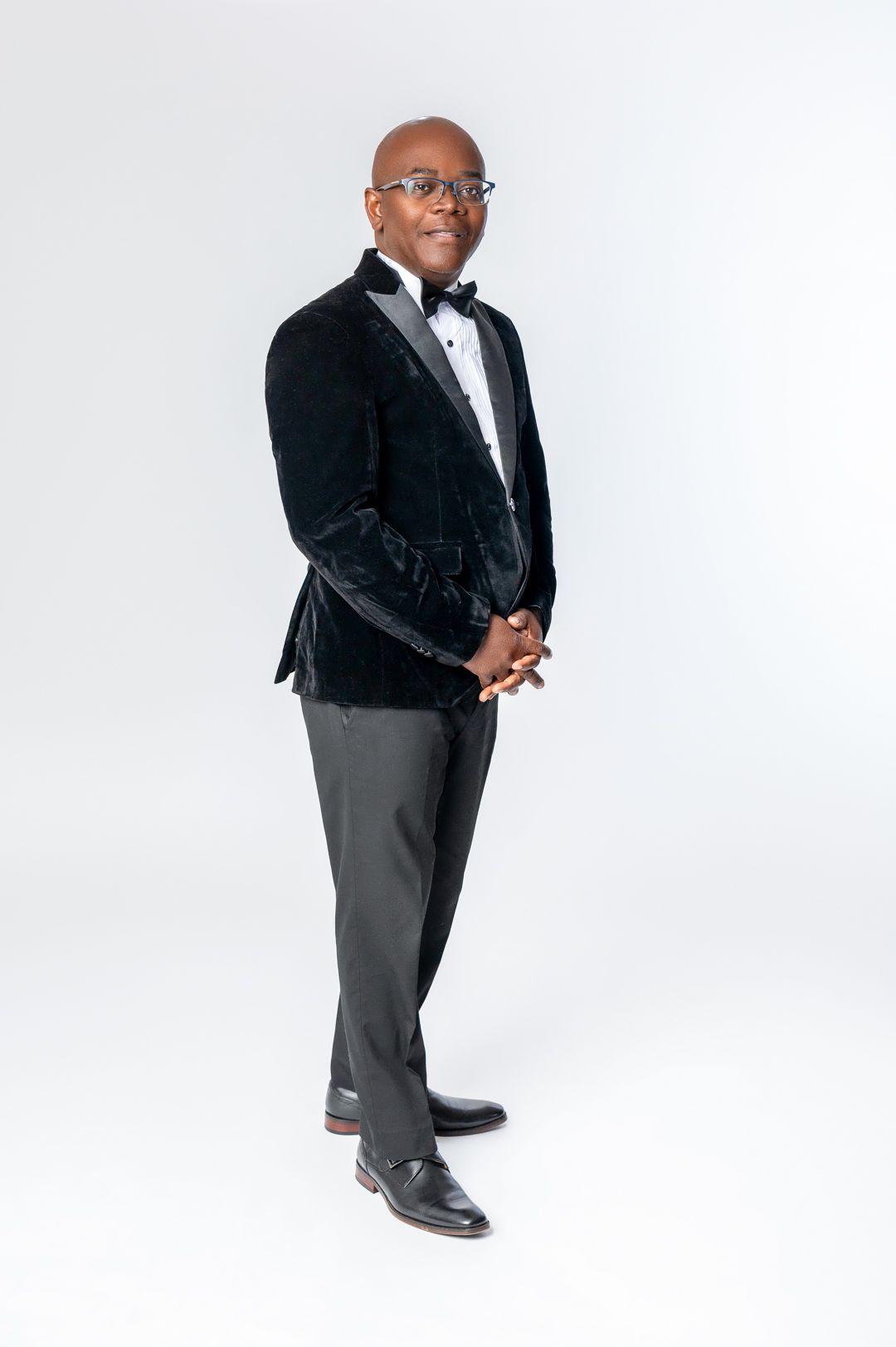 A formal portrait of someone in a black velvet tuxedo jacket and bow tie against white background.