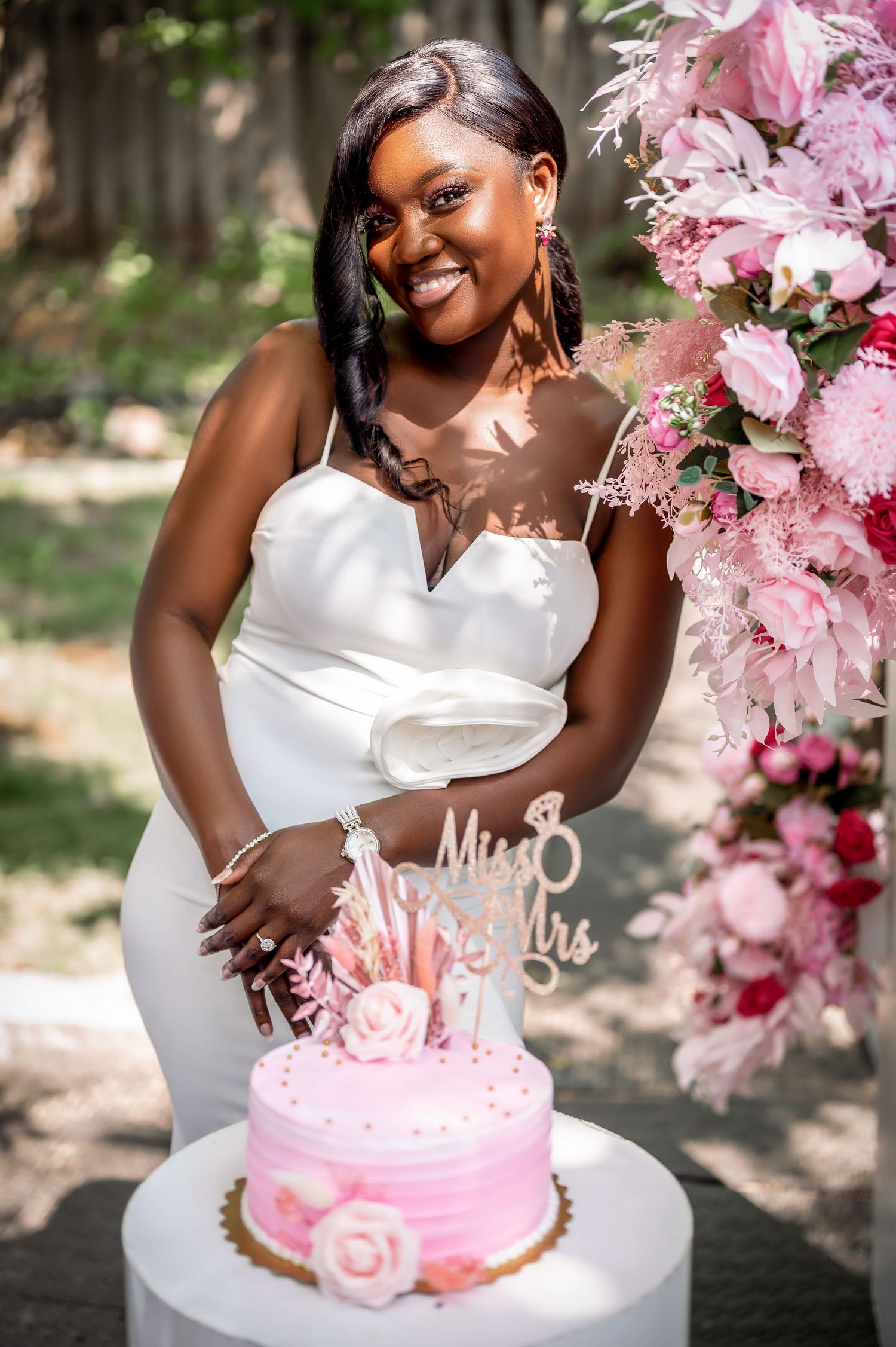 Bride poses with pink wedding cake decorated with roses in outdoor garden setting.