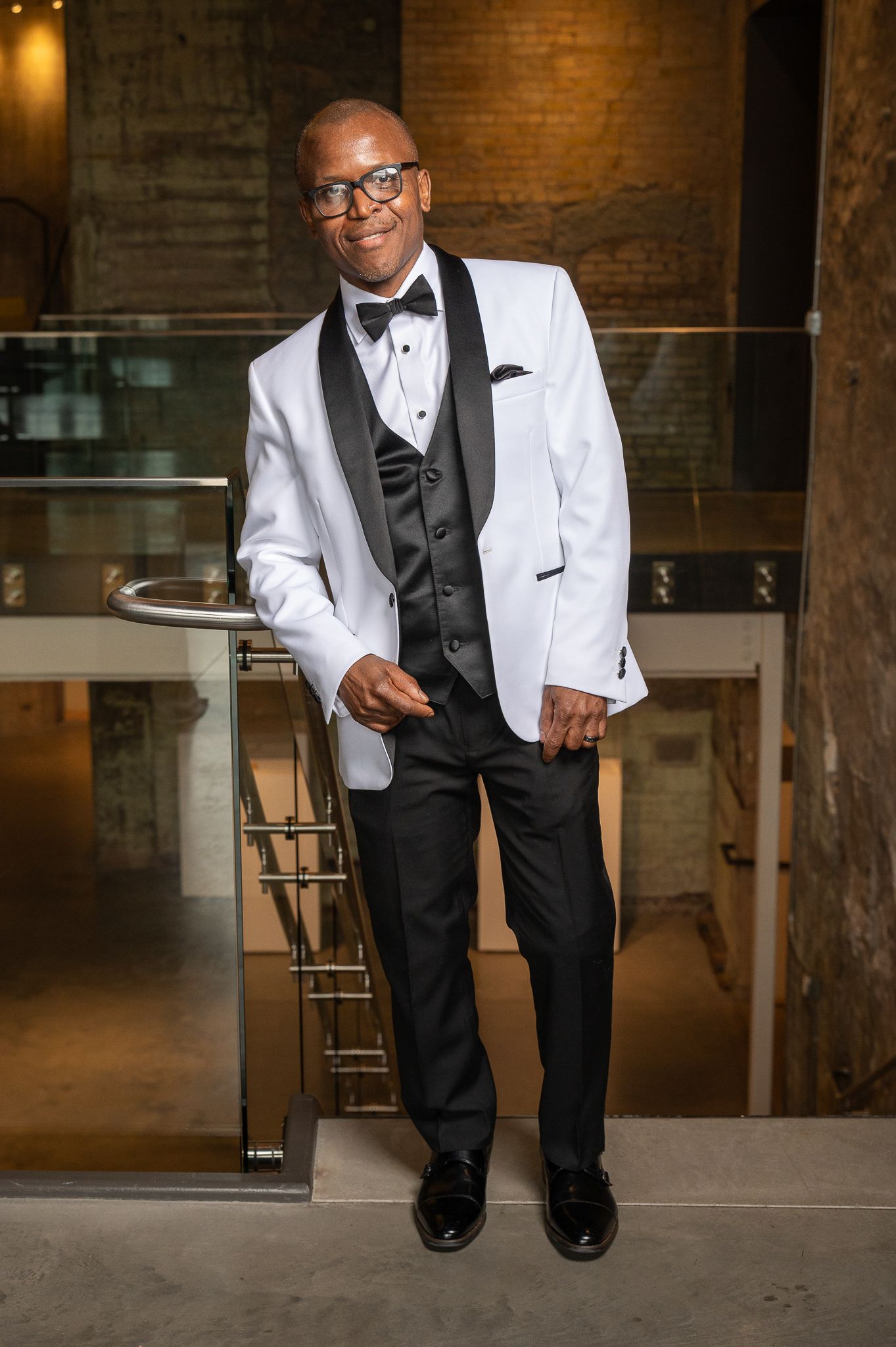 Groom in white tuxedo jacket with black bow tie and vest standing in modern venue.