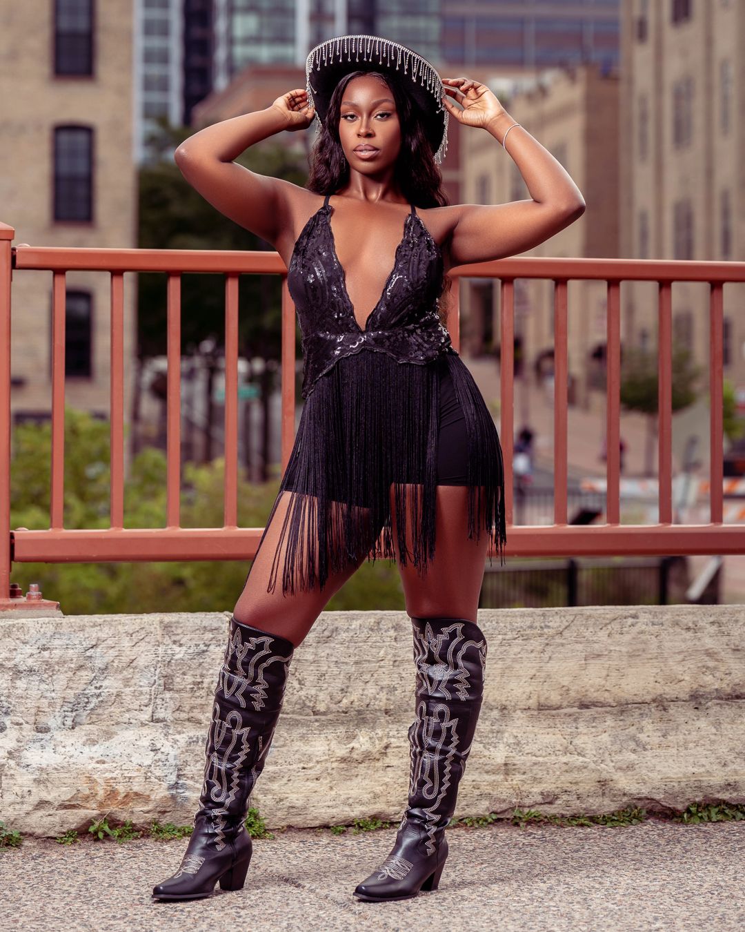 Urban fashion shoot with cowboy boots and fringe dress against industrial railing backdrop.