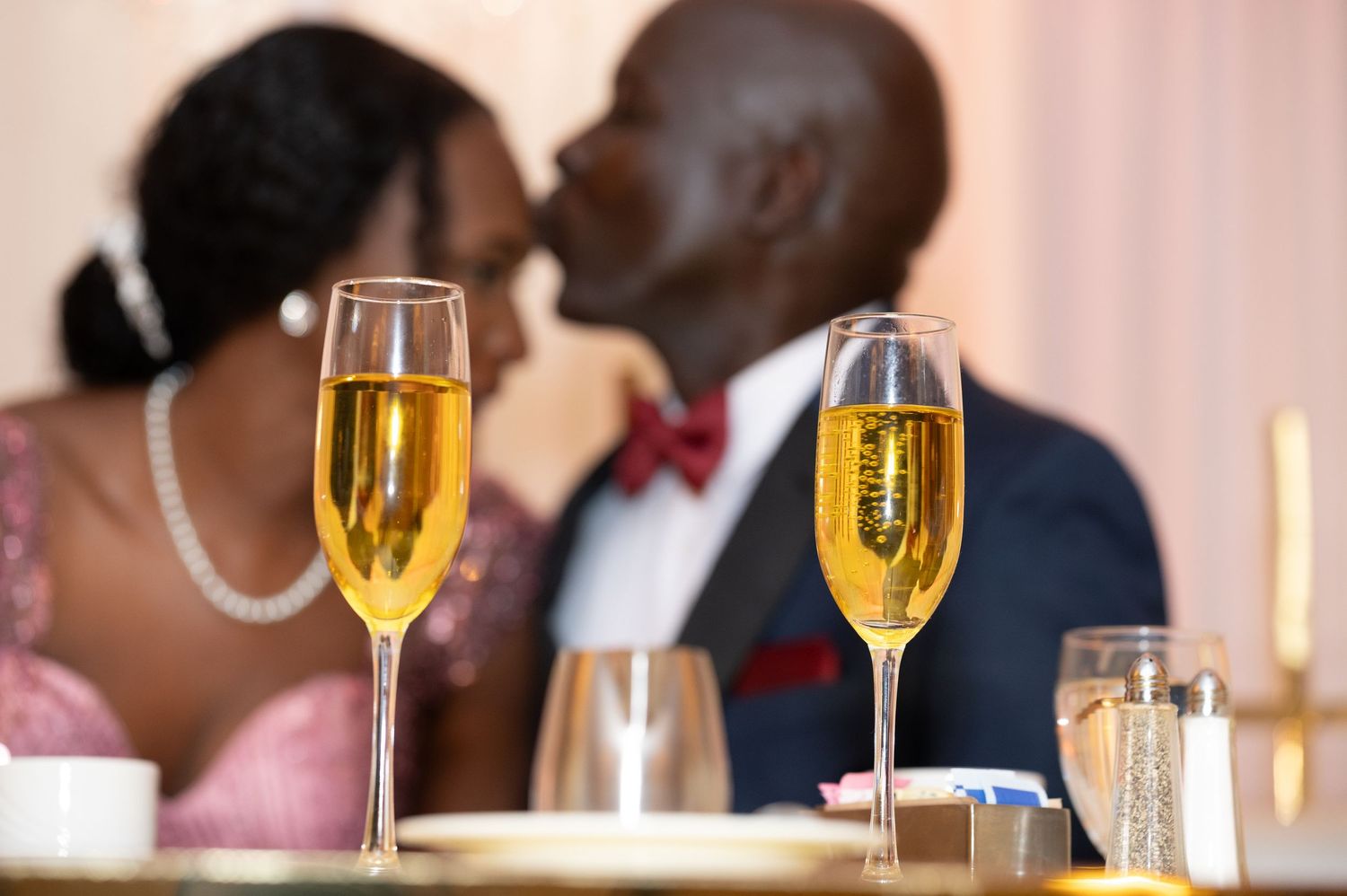Two champagne flutes on a wedding table with romantic couple sharing a kiss in the background.