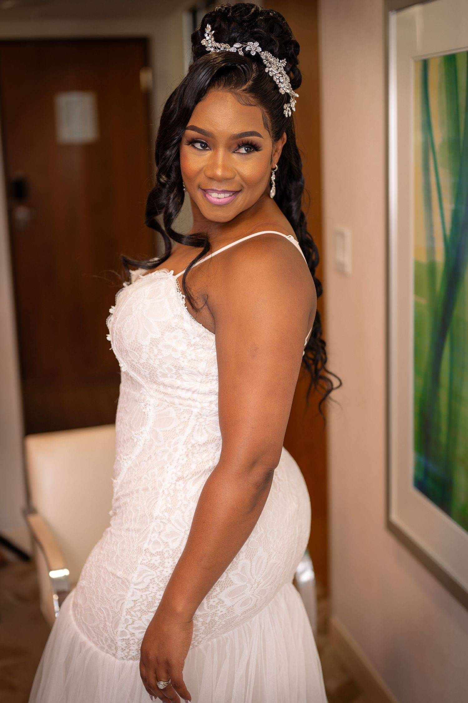 A person in a white lace wedding dress with crystal headpiece smiles elegantly.