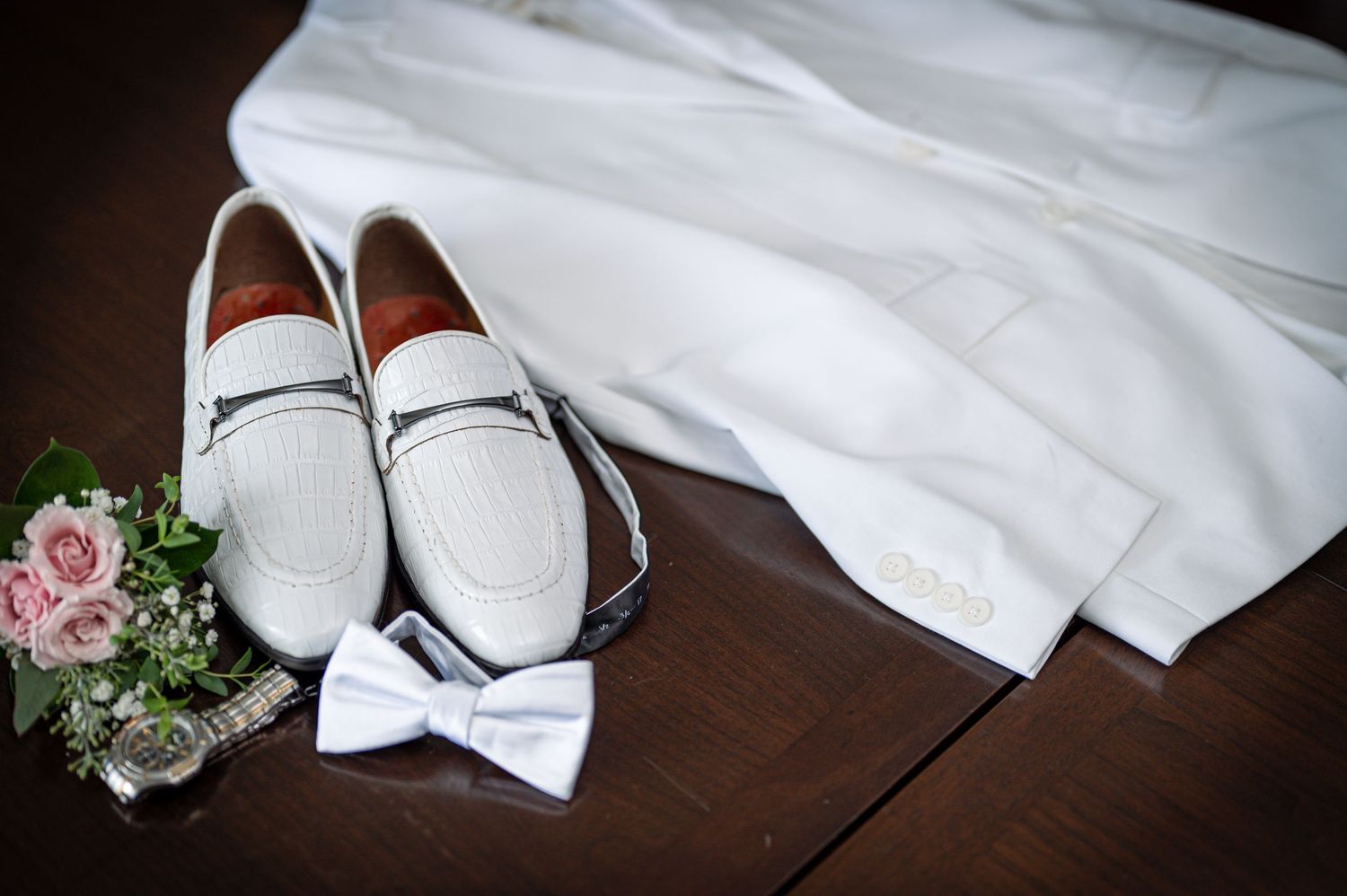 White formal leather loafers with a bowtie and small floral bouquet arranged on dark wooden surface.
